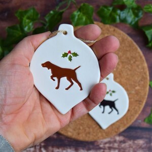 May include: White ceramic Christmas bauble shaped like a teardrop with a brown dog silhouette and a sprig of holly with red berries.
