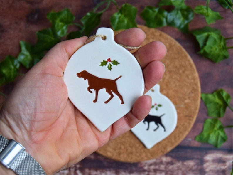 May include: White ceramic Christmas bauble shaped like a teardrop with a brown dog silhouette and a sprig of holly with red berries.