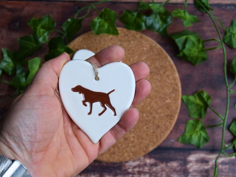 May include: White ceramic heart-shaped ornament featuring a brown silhouette of a German Shorthaired Pointer dog.