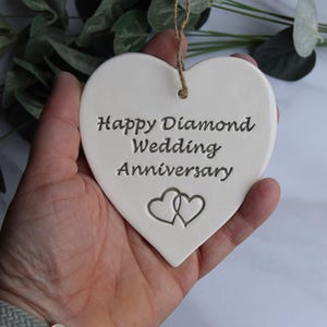 May include: A white heart-shaped ceramic ornament featuring the engraved text Happy Diamond Wedding Anniversary and two interlocking heart outlines. The ornament includes a brown twine string attached through a hole at the top for hanging.