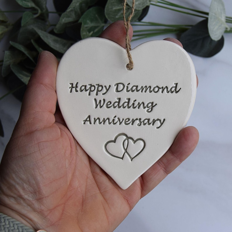 May include: A white heart-shaped ceramic ornament featuring the engraved text Happy Diamond Wedding Anniversary and two interlocking heart outlines. The ornament includes a brown twine string attached through a hole at the top for hanging.