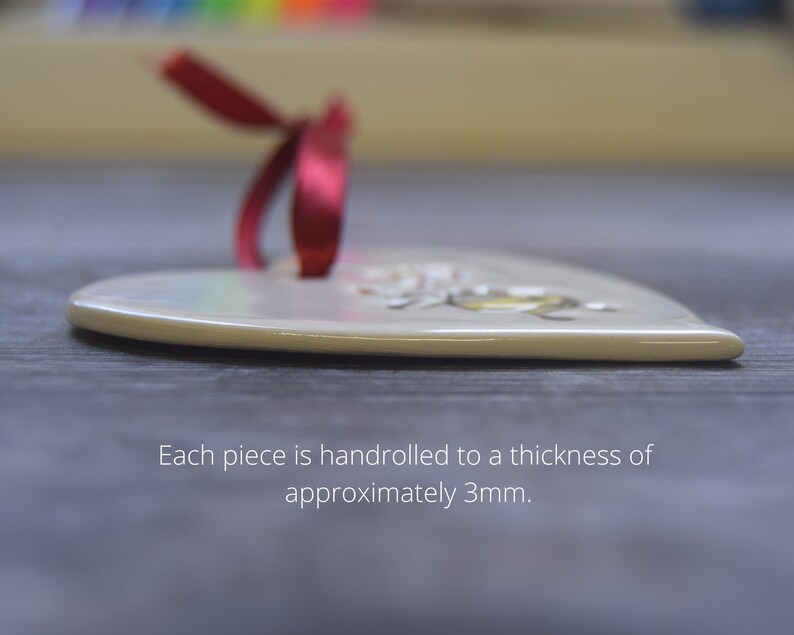 May include: A heart-shaped, cream-colored ornament with a red ribbon. The ornament has a floral design and is approximately 3mm thick. The text on the image reads: "Each piece is handrolled to a thickness of approximately 3mm."