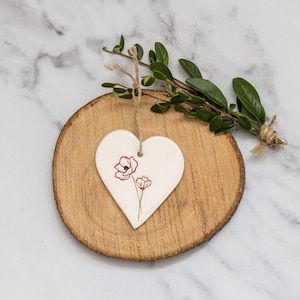May include: White ceramic heart-shaped ornament with a red poppy flower design on a wooden slice.