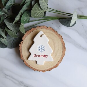 May include: A white ceramic Christmas tree ornament featuring a light blue snowflake graphic and the word Grumpy printed in red text. The ornament has a small hole at the top for hanging and is positioned on a circular natural wood slice.