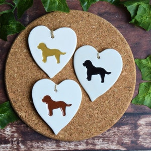 May include: Three white ceramic heart-shaped ornaments with dog silhouettes. The dogs are brown, black, and gold.