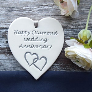 May include: A white heart-shaped ceramic plaque featuring the engraved text Happy Diamond wedding Anniversary and a design of two interlocking hearts. A small grey ribbon loop is attached to the top for hanging, centred above the text.