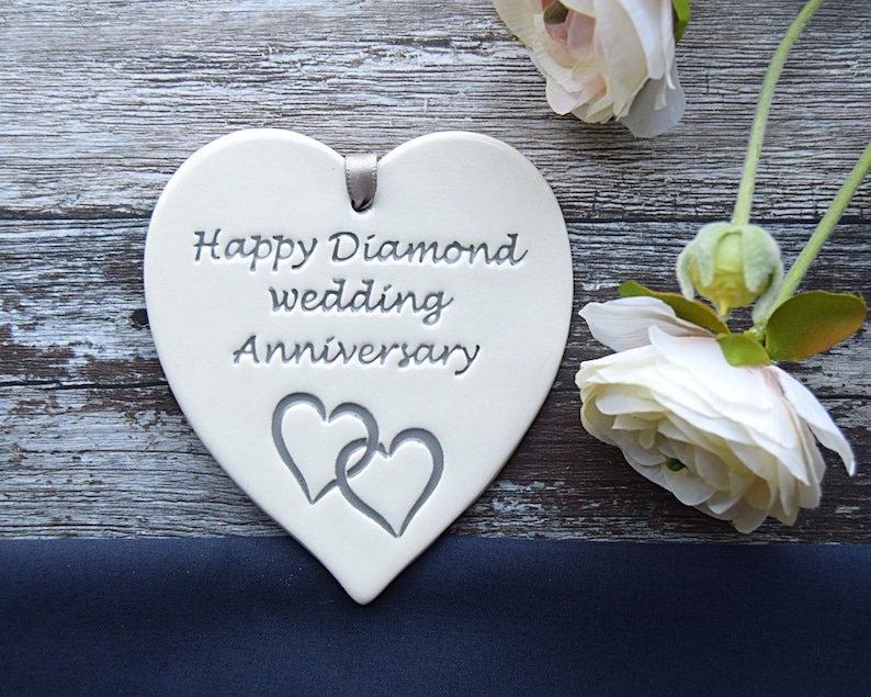 May include: A white heart-shaped ceramic plaque featuring the engraved text Happy Diamond wedding Anniversary and a design of two interlocking hearts. A small grey ribbon loop is attached to the top for hanging, centred above the text.