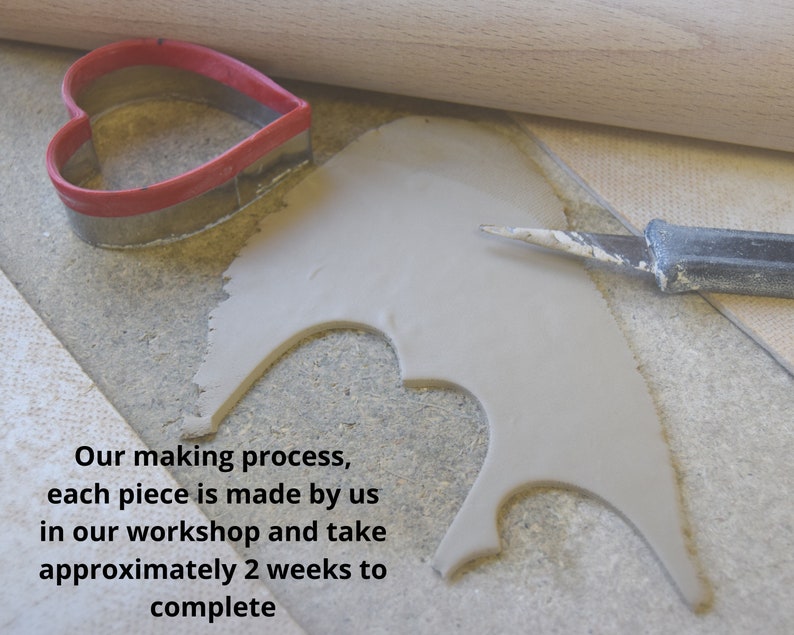 May include: A red heart-shaped cookie cutter, a wooden rolling pin, and a metal craft knife resting on a flat piece of grey clay. The text reads: Our making process, each piece is made by us in our workshop and take approximately 2 weeks to complete.