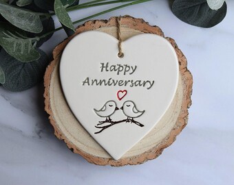 May include: A white heart-shaped ceramic plaque with the words "Happy Anniversary" and a red heart. Two birds are perched on a branch. The plaque is mounted on a wooden slice and has a jute string for hanging.