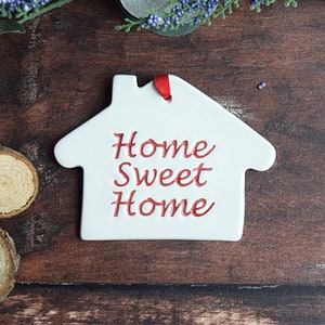 May include: White ceramic house-shaped ornament with a red ribbon and the text "Home Sweet Home" in red cursive lettering.