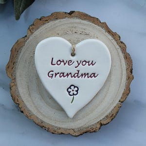 May include: A white heart-shaped ceramic ornament with the text "Love you Grandma" in burgundy script. A small flower is below the text. The ornament is on a round, natural wood slice with a bark edge.