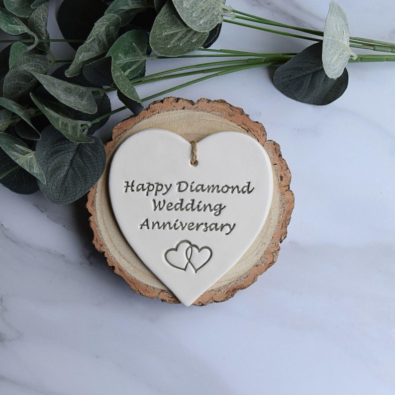 May include: A white heart-shaped ceramic ornament featuring the engraved text Happy Diamond Wedding Anniversary and two small interlocking heart outlines. The ornament hangs from a brown twine loop and rests on a circular natural wood slice.