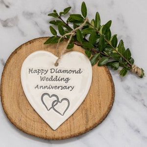 May include: A white heart-shaped ceramic ornament featuring the engraved text Happy Diamond Wedding Anniversary and two interlocking heart outlines. The ornament hangs from a natural jute twine string and is displayed on a round wood slice.