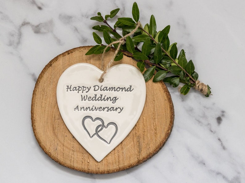 May include: A white heart-shaped ceramic ornament featuring the engraved text Happy Diamond Wedding Anniversary and two interlocking heart outlines. The ornament hangs from a natural jute twine string and is displayed on a round wood slice.