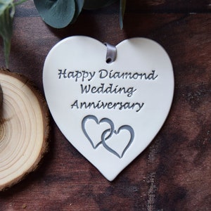 May include: A white heart-shaped ceramic ornament featuring the engraved text Happy Diamond Wedding Anniversary and a pair of interlocking hearts. A small grey ribbon is threaded through a hole at the top for hanging purposes.