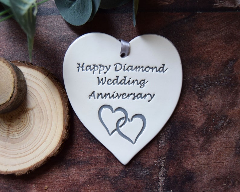 May include: A white heart-shaped ceramic ornament featuring the engraved text Happy Diamond Wedding Anniversary and a pair of interlocking hearts. A small grey ribbon is threaded through a hole at the top for hanging purposes.