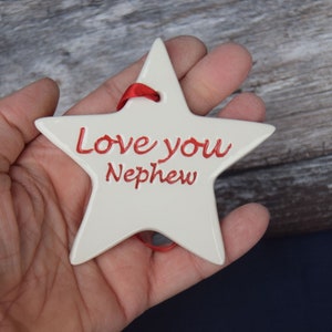 May include: White ceramic star ornament with a red ribbon. The star has the words "Love you Nephew" written in red.