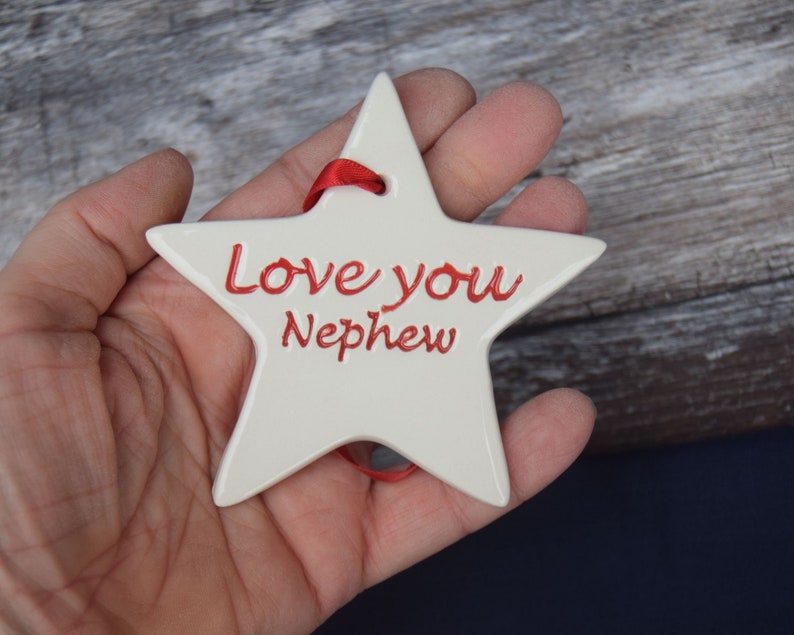 May include: White ceramic star ornament with a red ribbon. The star has the words "Love you Nephew" written in red.