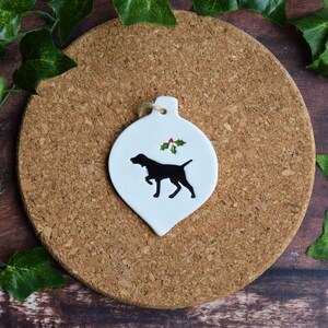 May include: White ceramic Christmas bauble shaped like a teardrop with a black silhouette of a dog and a green holly sprig with red berries.