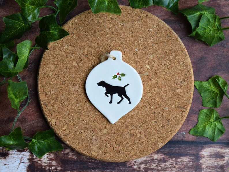 May include: White ceramic Christmas bauble shaped like a teardrop with a black silhouette of a dog and a green holly sprig with red berries.
