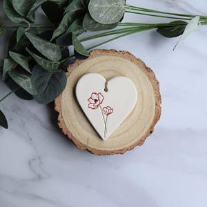 May include: A white ceramic heart-shaped ornament featuring a delicate red line drawing of two poppy flowers. The heart has a small hole at the top with a thin brown twine loop for hanging, resting on a circular slice of natural wood with visible bark.