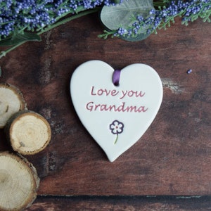 May include: A white heart-shaped ceramic plaque with the words "Love you Grandma" in red script. A small purple flower is below the text. A purple ribbon is attached at the top. The plaque is on a dark wooden surface with decorative elements.