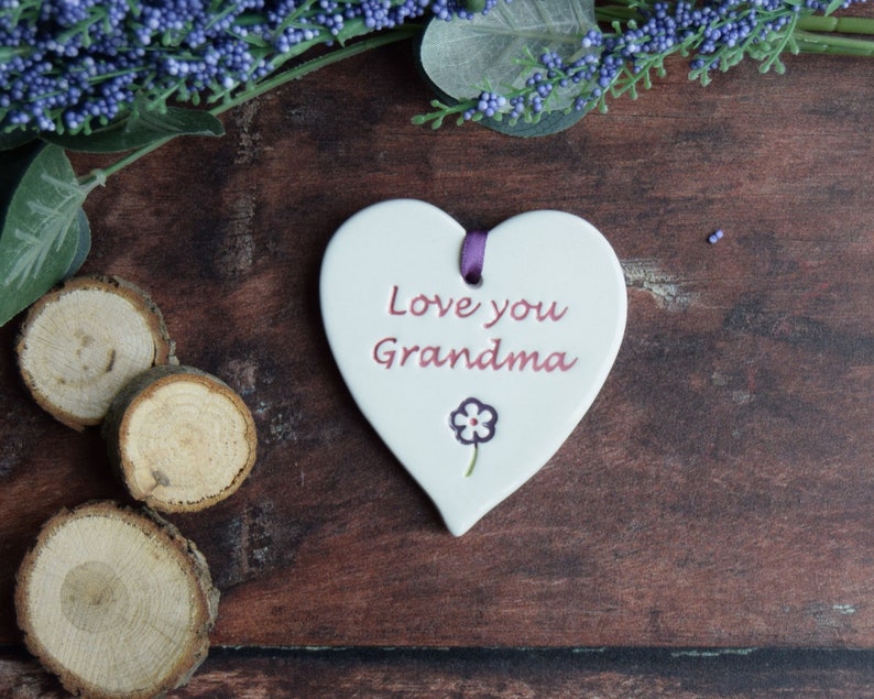 May include: A white heart-shaped ceramic plaque with the words "Love you Grandma" in red script. A small purple flower is below the text. A purple ribbon is attached at the top. The plaque is on a dark wooden surface with decorative elements.