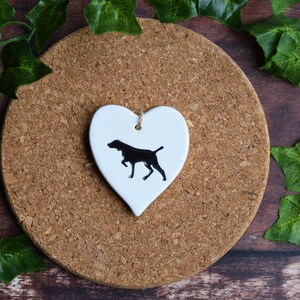 May include: White ceramic heart-shaped ornament with a black silhouette of a dog. The ornament is hanging from a string.