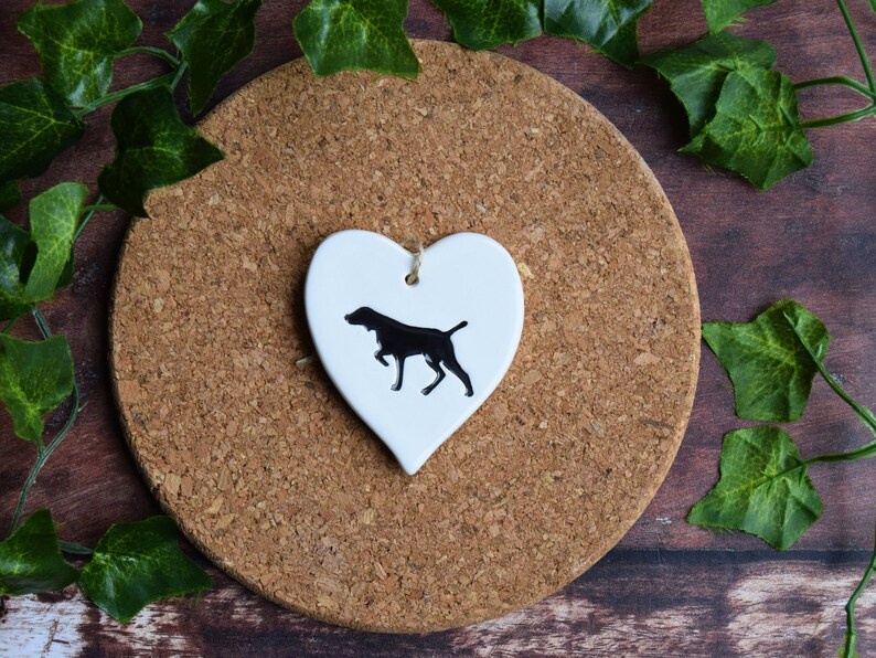 May include: White ceramic heart-shaped ornament with a black silhouette of a dog. The ornament is hanging from a string.