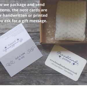 May include: A white cardboard box with a white tissue paper-filled pouch inside. The box is on a wooden surface. A business card with the text "Broadlands Pottery" and the website address "www.broadlandspottery.co.uk" is also on the surface. A folded card with the text "Just a note to say" is visible in the background.