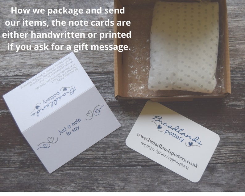 May include: A white cardboard box with a white tissue paper-filled pouch inside. The box is on a wooden surface. A business card with the text "Broadlands Pottery" and the website address "www.broadlandspottery.co.uk" is also on the surface. A folded card with the text "Just a note to say" is visible in the background.