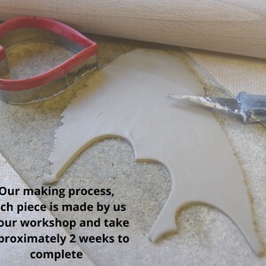 May include: A close-up of a grey clay piece being cut out with a small knife. A heart-shaped cookie cutter and a rolling pin are also visible. The text "Our making process, each piece is made by us in our workshop and take approximately 2 weeks to complete" is visible at the bottom of the image.