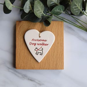 May include: White ceramic heart-shaped ornament with the text "Awesome Dog Walker" in red lettering and a black and white illustration of a dog on a leash. The ornament is displayed on a wooden surface.