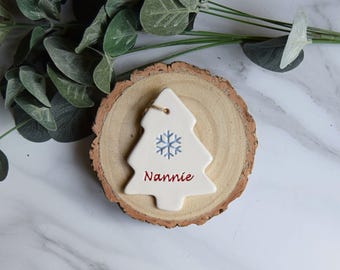 May include: A white ceramic Christmas tree ornament featuring a light blue snowflake graphic and the red text Nannie printed on the front. The ornament has a small hole at the top for hanging and is displayed on a circular piece of natural wood.