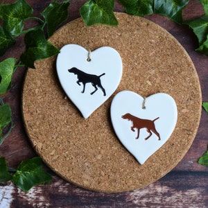 May include: Two white ceramic heart-shaped ornaments with black and brown dog silhouettes. The ornaments have a string for hanging.