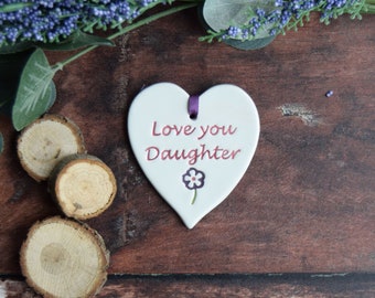 May include: White ceramic heart-shaped ornament with a purple ribbon and the text "Love you Daughter" in pink lettering. A small purple flower with a white center is below the text.