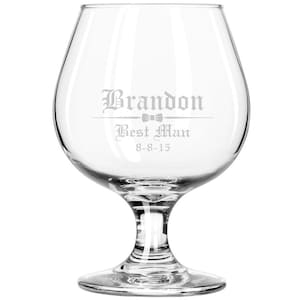 May include: A clear glass goblet with a stem and a round base. The goblet is engraved with the text "Brandon Best Man 8-8-15" and a bow tie.