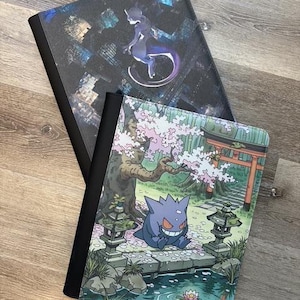 May include: Two rectangular binders with black spines. The top binder features a dark, city-themed background with a purple Mewtwo character. The bottom binder displays a Gengar character sitting on a stone bridge in a garden with cherry blossoms.