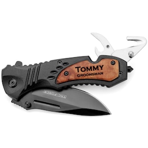 May include: A black folding pocket knife with a wood handle. The handle has the text "TOMMY GROOMSMAN" engraved on it. The knife has a bottle opener and a belt cutter.