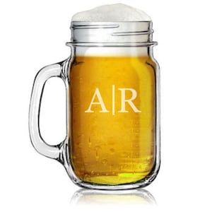 May include: A clear glass beer mug filled with golden beer and topped with white foam. The mug has a handle and the initials "AR" are etched on the front. The mug is sitting on a reflective surface.