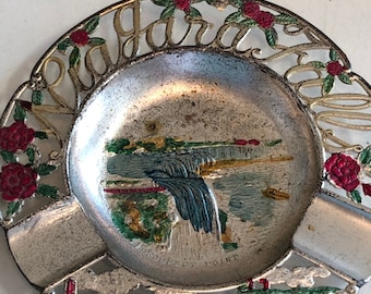 May include: A silver metal ashtray with a cut-out design of the word "Niagara Falls" around the edge. The center of the ashtray features a painted image of Niagara Falls with the words "Prospect Point" and "American Falls".
