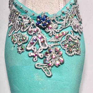 May include: A pair of turquoise ballet shoes decorated with silver lace and iridescent rhinestones. The shoes have a blue rhinestone flower accent.