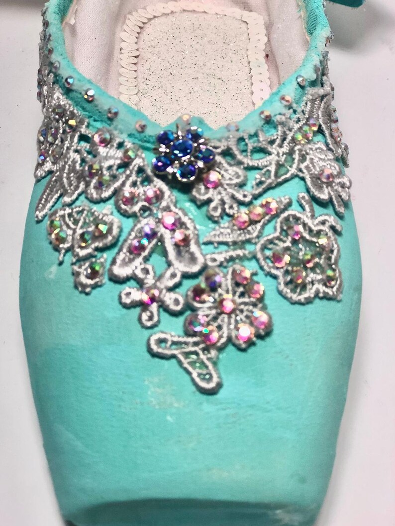 May include: A pair of turquoise ballet shoes decorated with silver lace and iridescent rhinestones. The shoes have a blue rhinestone flower accent.