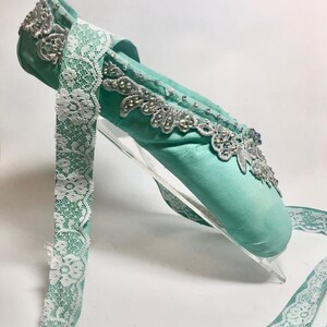 May include: A light blue ballet shoe with white lace trim and silver embellishments. The shoe is on a clear acrylic stand.