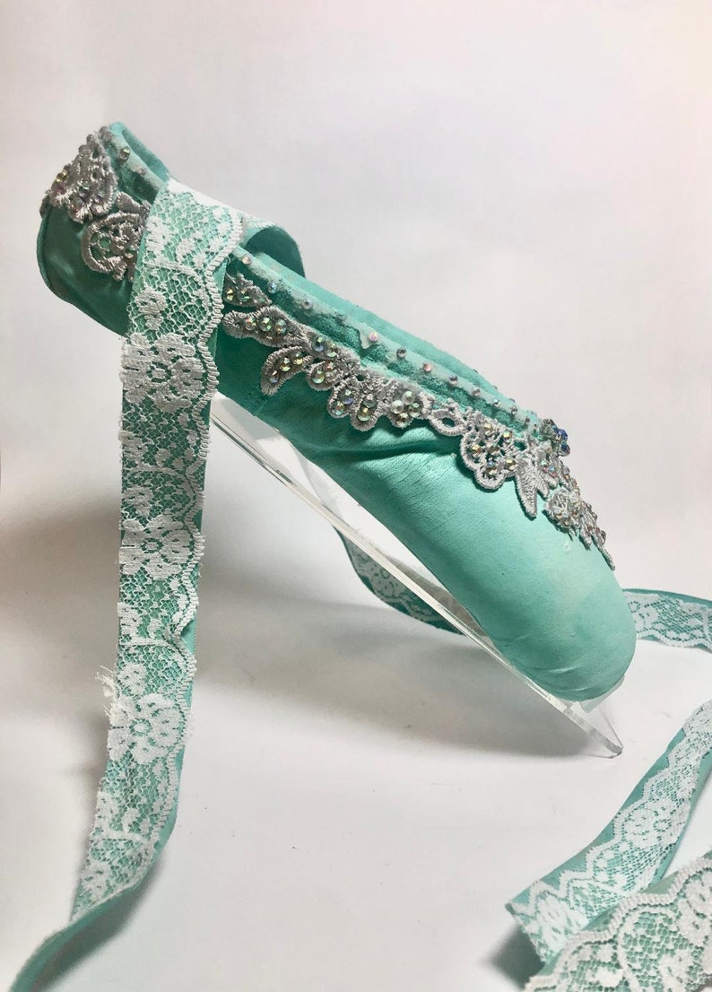 May include: A light blue ballet shoe with white lace trim and silver embellishments. The shoe is on a clear acrylic stand.