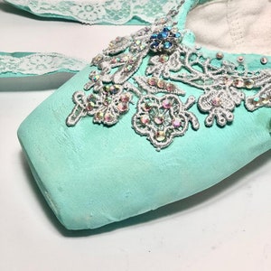 May include: A pair of light blue ballet shoes with a white lace ribbon and silver lace embellishments. The shoes are decorated with iridescent rhinestones and a blue jewel.