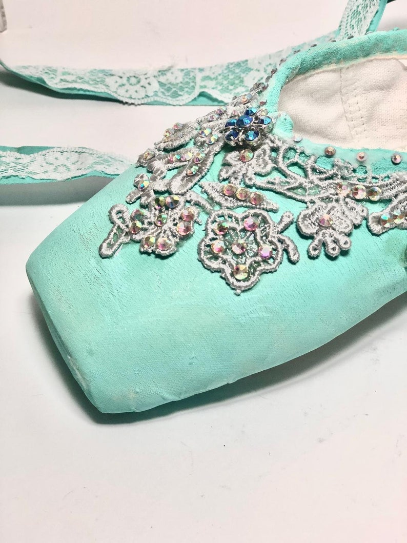May include: A pair of light blue ballet shoes with a white lace ribbon and silver lace embellishments. The shoes are decorated with iridescent rhinestones and a blue jewel.