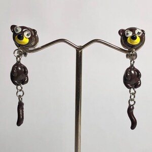 May include: A pair of brown bear dangle earrings with yellow eyes and a silver chain. The earrings are hanging from a silver stand.