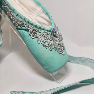 May include: A pair of light blue ballet shoes with a white lace trim and silver embellishments. The shoes are on a white background.