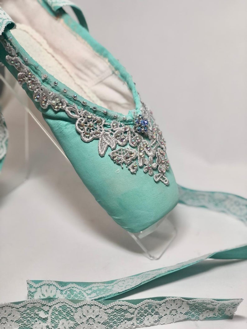 May include: A pair of light blue ballet shoes with a white lace trim and silver embellishments. The shoes are on a white background.